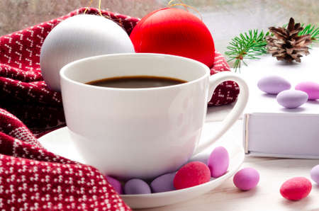 Cozy concept. Cup of coffee with colorful sweets, christmas toys and a warm scarf on a wooden windowsill against the background of a windowの写真素材