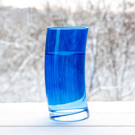 Deformed blue glass with clear spring water on the background of a snowy forestの写真素材