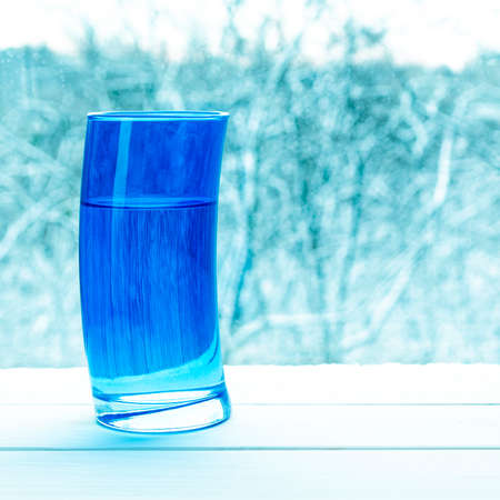 Deformed blue glass with clear spring water on the background of a snowy forestの写真素材