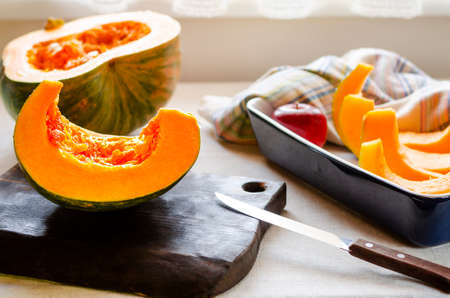 A bright orange pumpkin cut into pieces lies in a baking dish and on a chopping wooden board next to a knife and red applesの写真素材