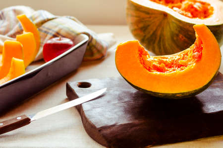 A bright orange pumpkin cut into pieces lies in a baking dish and on a chopping wooden board next to a knife and red applesの写真素材