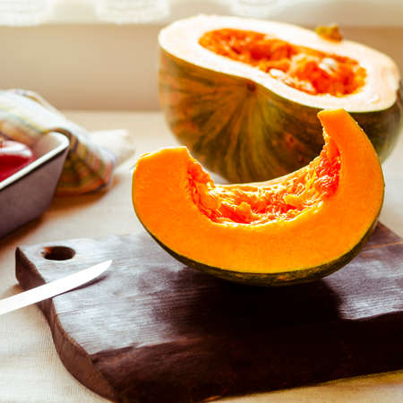 A bright orange pumpkin cut into pieces lies in a baking dish and on a chopping wooden board next to a knife and red applesの写真素材