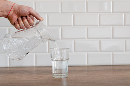 A jug of water in a man's hand. A glass of clean water on the kitchen table