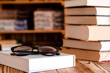 Book stacked on Laptop with glasses on wooden Table with blurred Bookshelf in library roomの写真素材