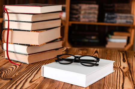 Black glasses lying on a white book next to old books against the background of bookshelvesの写真素材