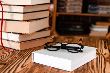Black glasses lying on a white book next to old books against the background of bookshelvesの写真素材