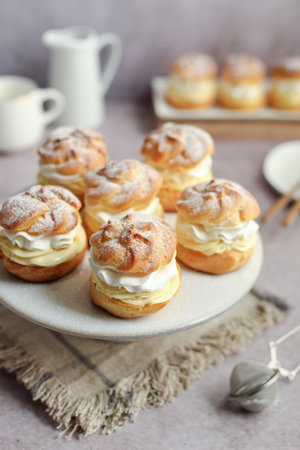 Cream puffs filled with vanilla cream and whipped cream on a white plate. Rustic napkin, white milk jug, and cup in the background.の写真素材