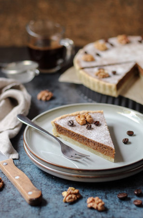 A slice of coffee and walnut tart on a grey plate. Homemade delicious dessert. Cup of coffee, blue base, and dark brown background.の写真素材