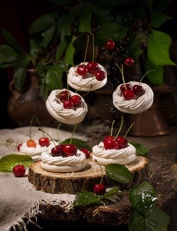 Delicate dessert with flying air meringue with natural cherries with cherry leaves on a tree cut, on a dark brown wooden table, on a dark chocolate background with plantsの写真素材