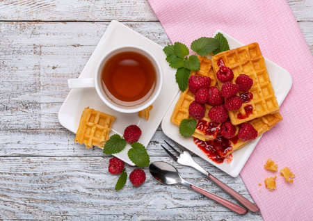 Viennese waffles with raspberries and raspberry jam, with mint leaves, lying on a white dish and a cup of green tea, top view on a light wooden table.の写真素材
