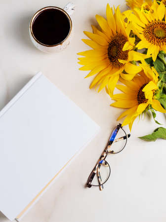 White book with space for text, coffee cup, yellow sunflower flowers on a marble background. Mockupsの写真素材