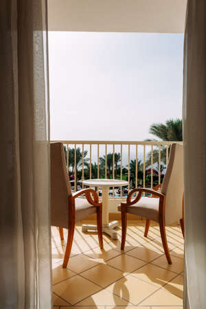 Two chairs and table on a balcony overlooking the Sea with grass and palm trees.