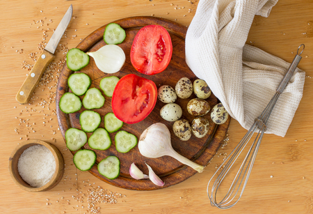 fresh cucumbers, tomatoes, onions, garlic, quail eggs, a knife, a towel on wood background.の写真素材