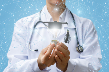 Doctor shows a light bulb in academic hat on blurred background.の写真素材