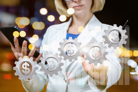 A businesswoman with a device in her hand is showing a cogwheels system with the currency unit inside. The concept is the analytics and successful financial management.の写真素材