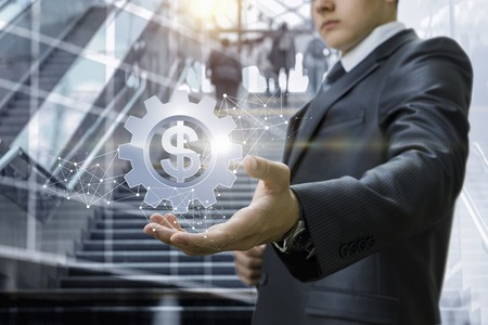 The concept is profitable investments. A businessman is holding a cogwheel with currency unit inside surrounded by wireless connections at the busy commercial centre background.の写真素材