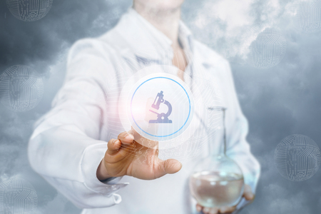 A laboratorian with a sample flask in one hand is touching the microscope icon on the touchscreen at the cloud sky background. The concept is the laboratorian researchers.の写真素材