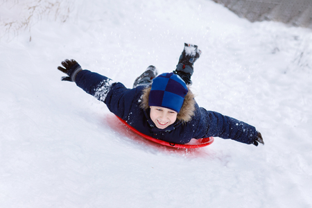 A boy is enjoying sledging laying on the red plastic plate with his hands moving apart from the huge snow hill. The concept is children's winter outdoor activity.の写真素材