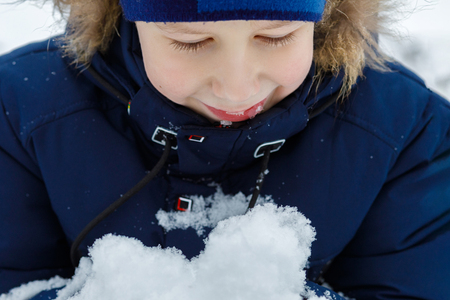 A happy, smiling boy is looking at the snow heap in his hand enjoying the outdoor winter pastime. The concept is children playing in winter.の写真素材