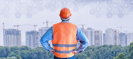 An engineer in the uniform is standing with his back and looking at the skyscrapers construction...