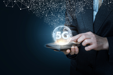A businessman in suit is holding a device in his hands with a global map with large 5G symbol on it illuminating a cobweb of network public connections at dark background.の写真素材