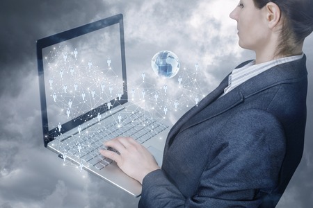 A businesswoman is standing half turn operating with notebook with networking scheme consisting of connections, human figures icons and a global map. The concept of global social networking system.の写真素材