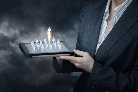 A businesswoman is holding a device with human figures icons standing on it with one of them raising up at grey background. The concept of choice, challenge and solution.の写真素材
