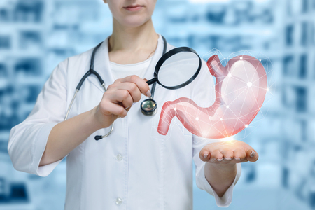 A young doctor with a stethoscope around her neck is standing and looking at the stomach model hanging above her hand through a loupe at the blurred laboratorian room background.の写真素材
