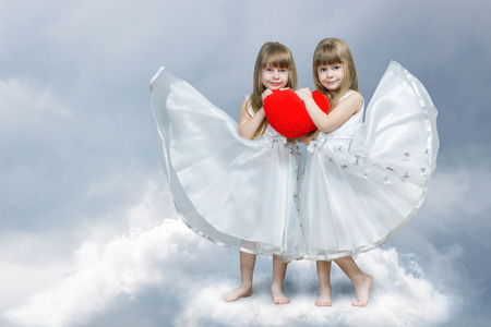 Two smiling cute blondie sisters twins in white dresses are holding a big bright red heart together standing barefoot on the cloud at light blurred background. The concept of tight family relations.の写真素材