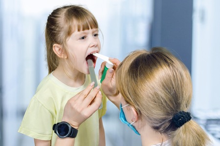 A closeup of female pediatrician treating a throat of a small child with widely opened mouth using a...