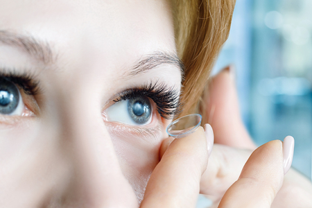 A closeup of a young woman inserting a transparent contact lens into her eye at blurred hospital room background. The concept of ophthalmological poor sight correction.の写真素材
