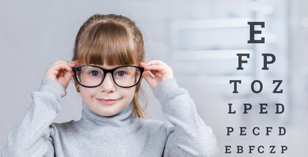 A smiling child in glasses is standing with checking table with letters behind her at the blurred...