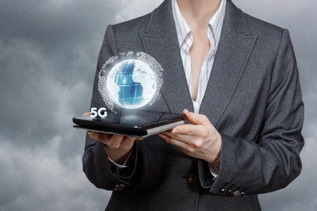 A closeup of a businesswoman holding a gadget with globe and 5G symbol hanging above it at grey sky background. Global high speed internet concept.の写真素材