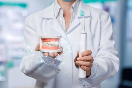 A closeup of a dentist showing an artificial jaw model and electric toothbrush at the blurred dental office background. The teeth hygiene concept.の写真素材