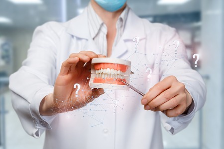 A closeup of a doctor holding a denture model surrounded with wireless connections and question marks and pointing at teeth with some medical tool. The concept of proper diagnostics and treatment.の写真素材