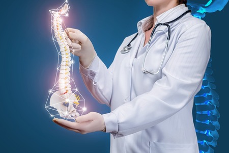A closeup of a physiotherapist with a stethoscope operating with a spine model with pelvis element with at dark background with digital spine image. A concept of spine diseases treatment.の写真素材