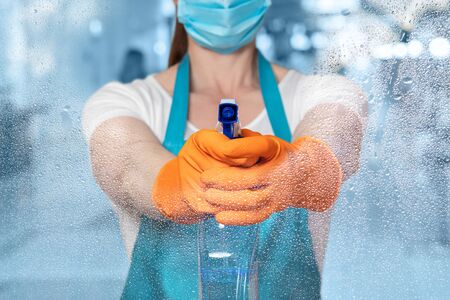 A cleaning lady in a mask is aiming a spray on a blurred background.
