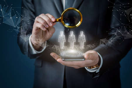 Business man examine through a magnifier a hologram of workers on a mobile device.の写真素材