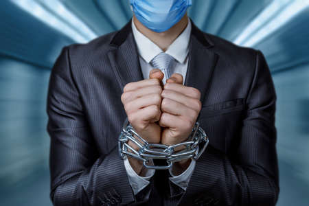 Concept of businessman problem due to coronavirus. Businessman behind forged hands in chains in a mask on a blurred background.の写真素材