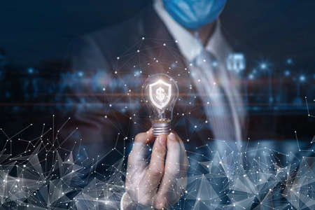 Businessman shows a light bulb with a burning shield and a dollar inside on a blurred background.