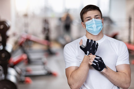Masked athlete wears fitness gloves on the background of the gym.の写真素材