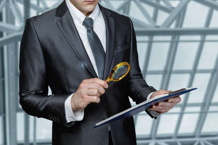 Young man looking through a magnifying glass at a document on a blurred background.の写真素材