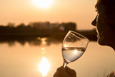 Woman looking into the distance with a glass of wine against the background of a sunny sunset river.の写真素材