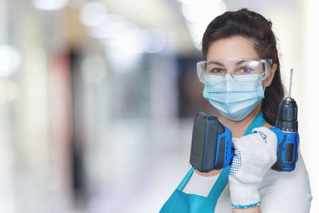 Female worker with a drill in her hands and a protective mask and goggles on a blurred background.の写真素材