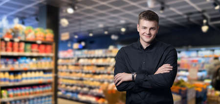 A young businessman in a black shirt standing with his arms crossed against the backdrop of a supermarket.の写真素材