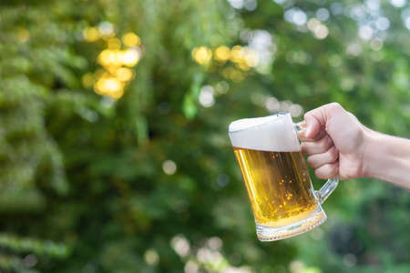 A glass of beer in a hand on a blurred background of trees.の写真素材