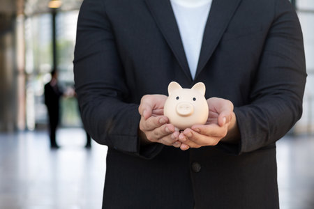Concept of savings in business. A businessman shows a piggy bank against the backdrop of the Hall of the business center.の写真素材