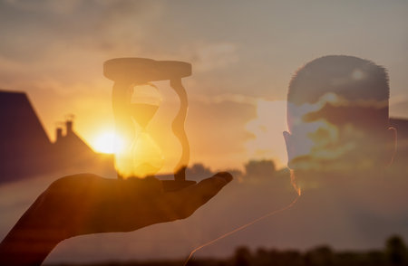 Double exposure of an hourglass in hand and a businessman looking into the distance against the backdrop of a sunset.の写真素材