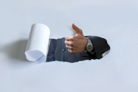 The concept of approval or success. A hand in a suit making a thumbs up gesture breaks through a sheet of paper.の写真素材