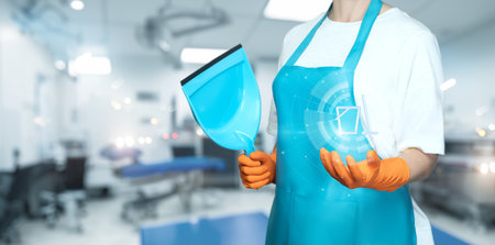 A professional cleaning in an apron holds a scoop and a digital device in a hospital setting. The individual is focused on maintaining cleanliness in a medical environment.の写真素材